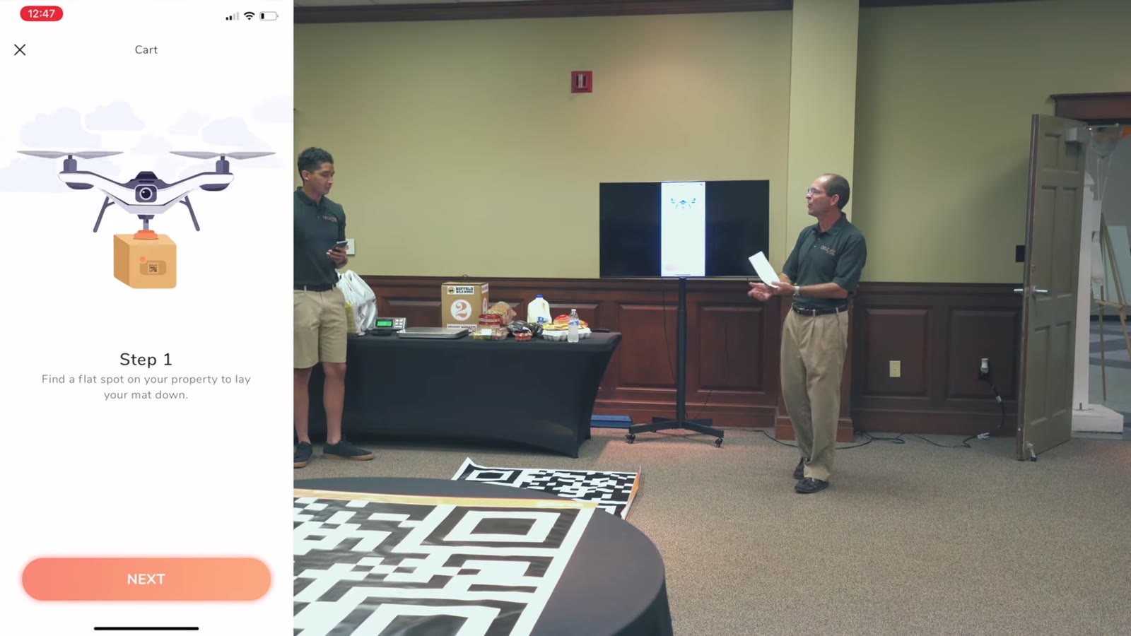 Two men demoing in a conference room, a QR landing mat on the floor and a Buffalo Wild Wings box on the table, with the Deuce Drone app picture-in-picture showing a drone illustration, Step 1, and an orange Next button