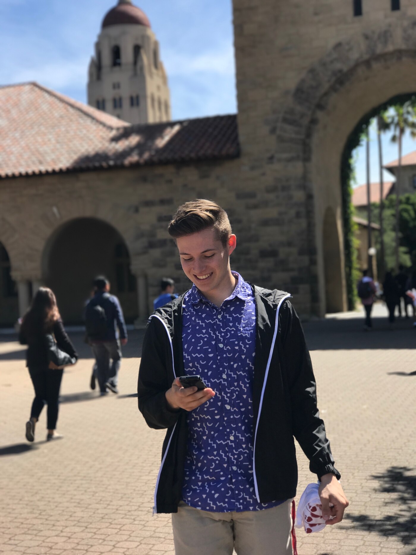 A teenager in a patterned shirt and windbreaker walking through a Stanford archway, smiling at his phone, Hoover Tower behind him