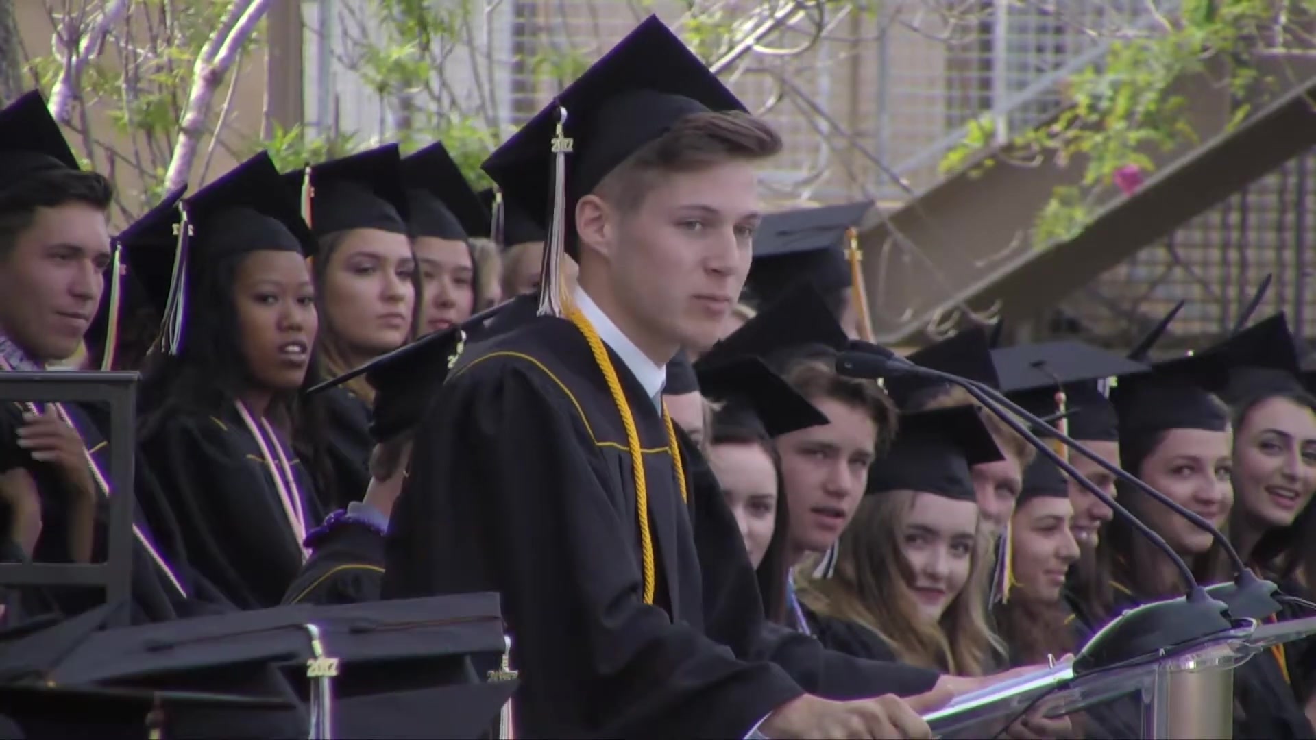 A graduate in cap and gown at the podium, seen from the side, looking up at the audience with a slight smile, rows of classmates in caps and gowns behind him