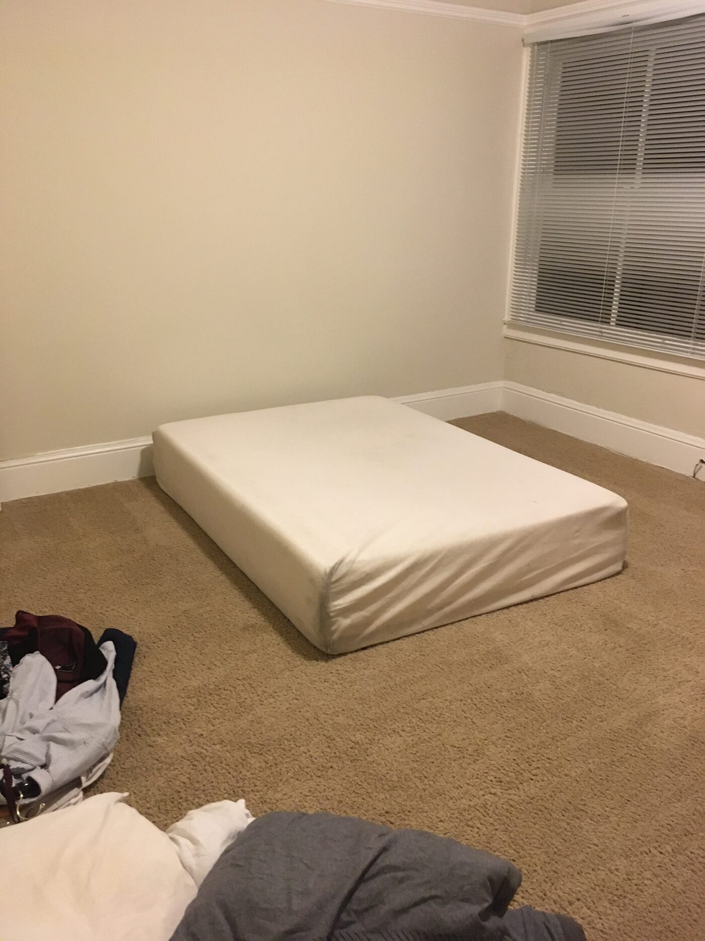 A bare mattress on the carpet of an otherwise empty room, clothes piled beside it
