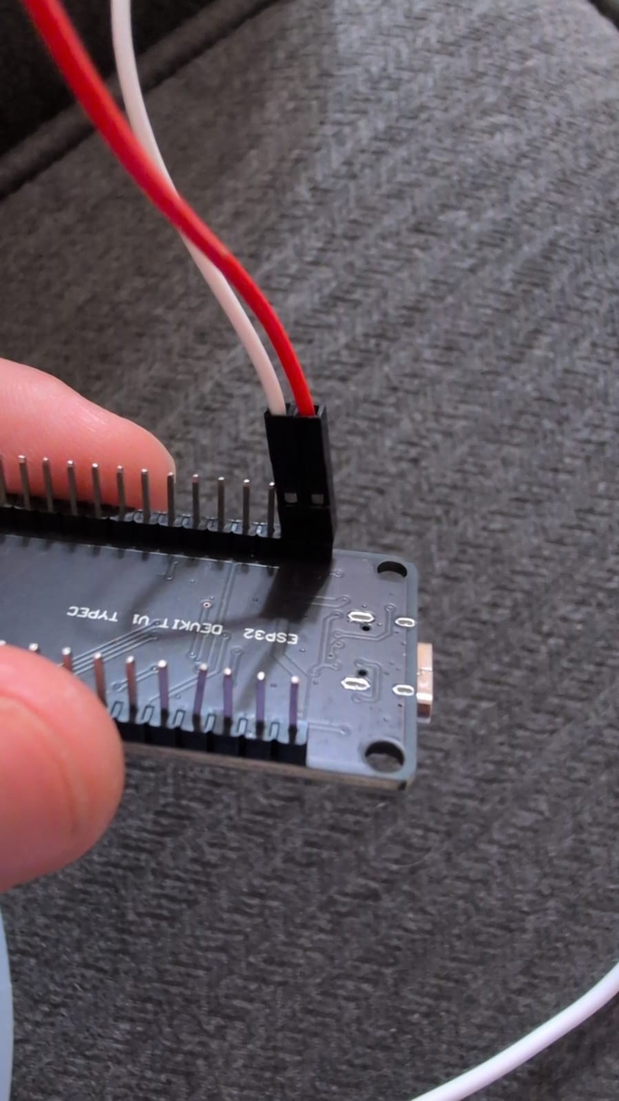 A hand holding a small dark circuit board with ESP32 DEVKIT V1 TYPEC silkscreened on the back, two jumper wires plugged into the header pins